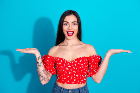 Smiling young woman in a red floral top gestures cheerfully against a vibrant blue background, showcasing trendy style and lively characterの写真素材
