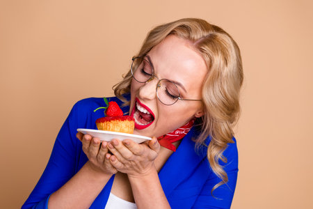 Elegant woman with blonde curls enjoying a strawberry dessert while smiling against beige backgroundの写真素材