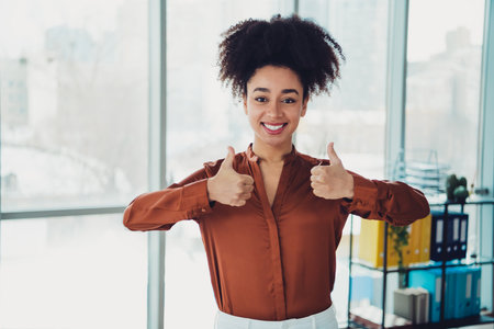 Smiling young professional woman giving thumbs up gesture in a stylish modern office with bright natural lighting and a cozy interiorの写真素材