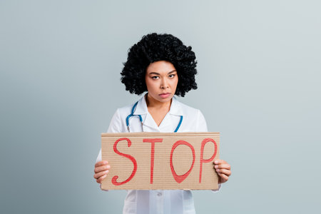 Serious female doctor holding a stop sign against a plain gray background, highlighting awareness and healthcare issuesの写真素材
