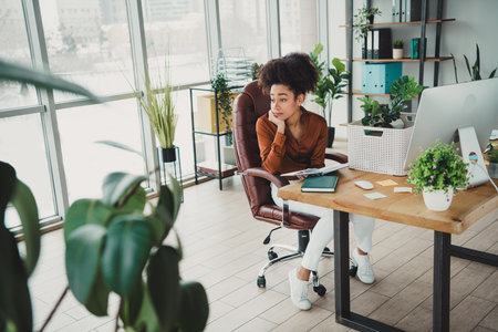 Young businesswoman sitting in a modern office with houseplants, thoughtfully glancing aside on a bright dayの写真素材