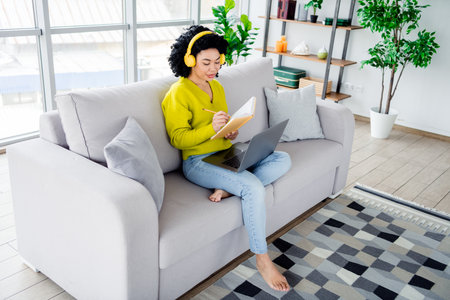 Young woman sitting on a sofa in a modern living room, wearing headphones and writing in a notebook with a laptop on her lapの写真素材