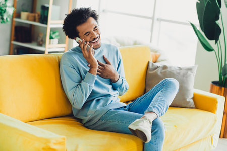 Young man on a yellow couch enjoying a cheerful phone conversation, radiating happiness and comfort in a cozy homeの写真素材