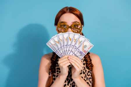 Young red-haired woman in stylish attire with glasses holding cash, standing against a blue backgroundの写真素材