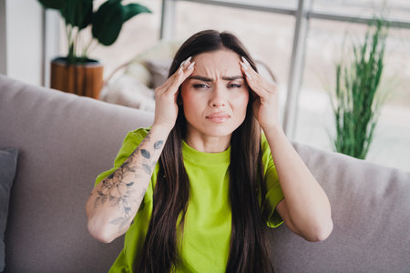 Young woman with brown hair indoors expressing emotion while sitting on a comfortable sofa in a bright home interiorの写真素材