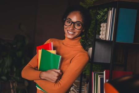 Young woman in casual orange turtleneck holding colorful notebooks, smiling confidently indoors, highlighting a cozy study atmosphere.の写真素材