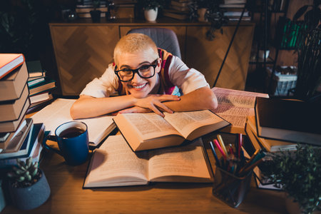 Young woman with glasses and short blond hair smiling amidst open books, pens, and a mug, studying thoughtfully in a warmの写真素材