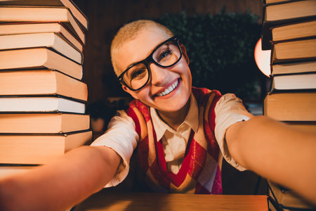 Young woman with glasses and short blond hair smiling indoors, surrounded by stacks of books, showcasing a casual academic styleの写真素材