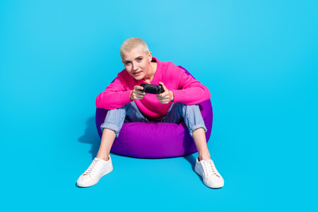 Joyful young woman in a trendy pink sweater sitting on a purple bean bag, holding a game controller...