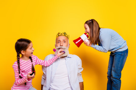 Grandfather Wearing a Crown Interacting Joyfully with Two Granddaughters in Front of Yellow Backgroundの写真素材