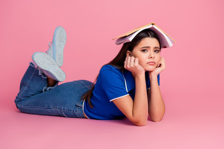 Young woman in casual outfit looking upset with book on her head on a pink background