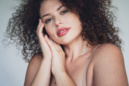 Confident young woman with curly brown hair embracing natural beauty against a simple backgroundの写真素材