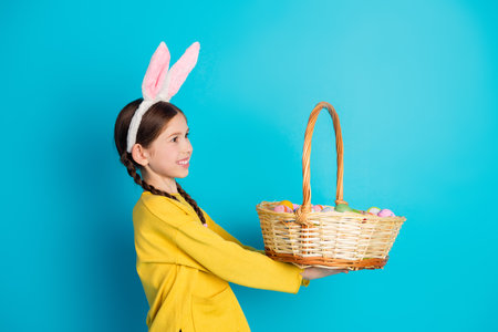 Adorable girl with bunny ears holding a wicker basket of painted Easter eggs against a bright blue...