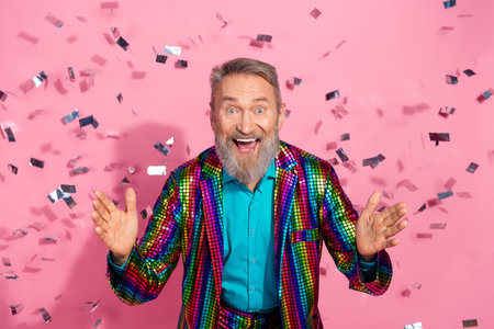 Cheerful elderly man in vibrant disco suit celebrating with confetti against a pink backgroundの写真素材