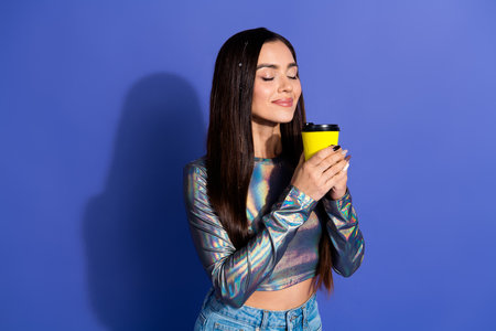 Young woman with holographic top savoring a coffee against a purple backdrop, showcasing modern style and relaxed enjoymentの写真素材