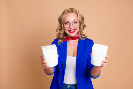 Cheerful woman in blue jacket showcasing white containers against a beige backgroundの写真素材