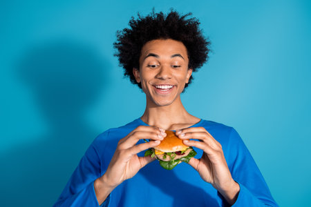 Photo portrait of handsome young guy eat burger bite dressed stylish garment isolated on blue color backgroundの写真素材