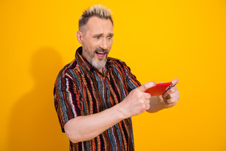 Charismatic mature man holding smartphone with humorous expression on vibrant yellow background wearing fashionable printed shirtの写真素材