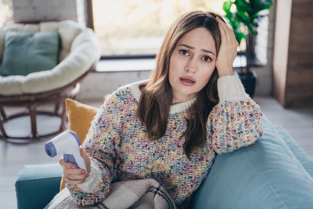 Young woman holding thermometer while sitting at home looking concerned