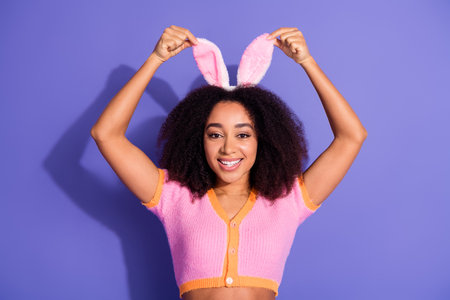 Smiling young woman with curly hair wearing bunny ears and posing against a purple background, exuding happy vibesの写真素材