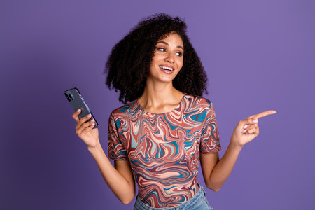 Stylish young woman with curly hair wearing colorful t-shirt poses with mobile phone against vibrant...