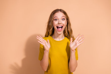 Excited young woman expressing surprise and wearing a fashionable yellow shirt against a beige backgroundの写真素材