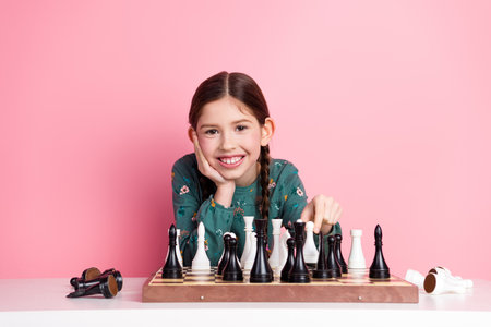 Young girl with charming smile playing chess on a pink background, showcasing joy and intelligence while enjoying leisureの写真素材