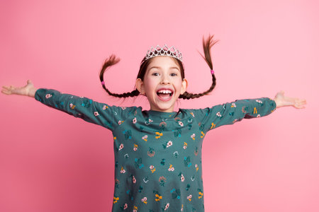 Smiling young girl wearing a crown with braided hair extending arms joyfully against a pink background in a stylish outfitの写真素材