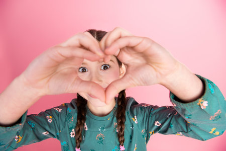 Young girl creating a heart shape with her hands against a pink background, expressing joy and fun...