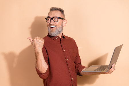Smiling mature man holding a laptop and showing a thumb gesture against a beige backgroundの写真素材