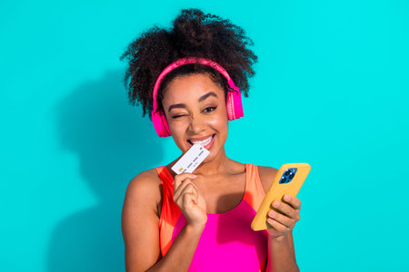Smiling young woman wearing headphones holding a credit card and phone against a turquoise...