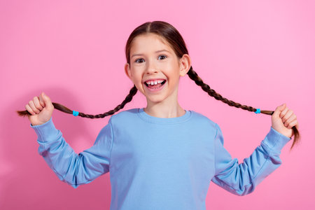 Cheerful young girl with braided hair in a blue sweatshirt smiling joyfully against a bright pink...