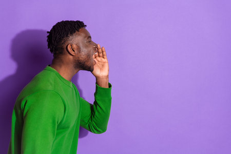 Young man shouting with hand near mouth against vibrant purple background, wearing green sweatshirt representing casual styleの写真素材