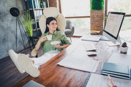 Young businesswoman relaxing at her office desk with coffee in hand, wearing elegant blouse, embodying success and professionalismの写真素材