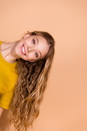 Portrait of a smiling young woman with wavy hair posing against a beige vertical backdrop in a...