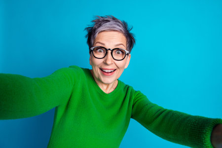 Joyful senior woman with glasses taking selfie against vibrant blue backgroundの写真素材