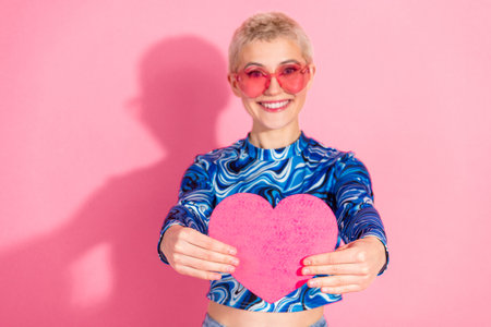 Smiling young woman with short blonde hair holding a pink heart on a vibrant background showcasing trendy fashion styleの写真素材