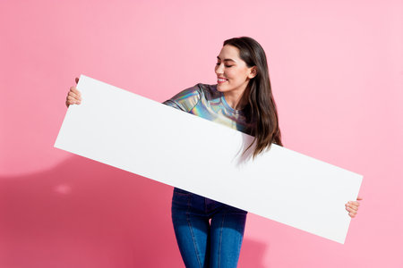 Young woman in holographic top holds white rectangular banner with a cheerful smile against a pink backgroundの写真素材