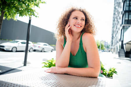 Young woman enjoying sunny day outdoors, sitting at an urban cafÃ© table. She is smiling brightly and looking into the distanceの写真素材