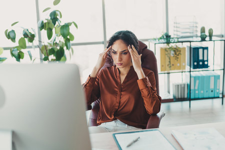 Young female professional experiencing workplace stress in a modern office environment with natural lighting and contemporary decor.の写真素材