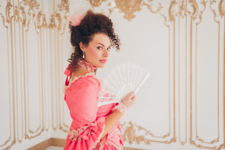 Elegant young woman in vintage pink gown holding a lace fan indoors