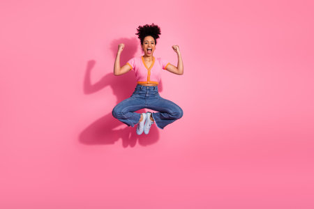 Confident young woman jumping with enjoyment in casual outfit against pink background expressing...