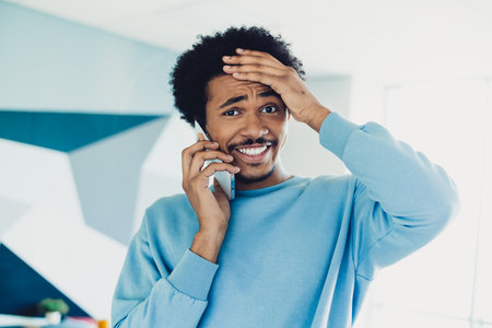 Young man expressing surprise while talking on the phone in a casual blue sweatshirt indoors,...