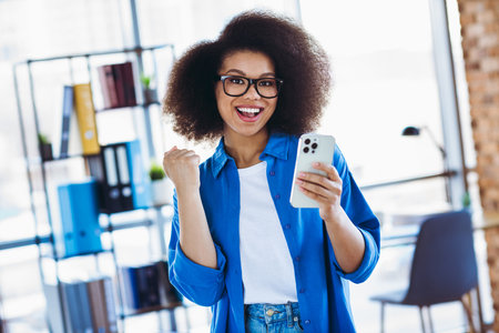 Cheerful young professional woman with smartphone celebrating success in a modern stylish office...