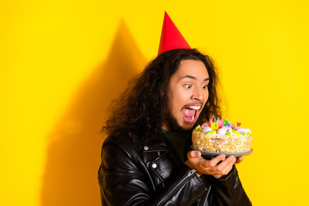 Young man celebrating his birthday with a cake, wearing a party hat against a vibrant yellow...