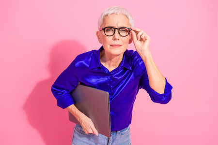 Confident mature businesswoman holding laptop and adjusting glasses on pink background