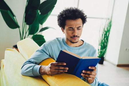 Young man reading a book on a yellow sofa in a modern bright interior, wearing a blue sweatshirt and enjoying leisure timeの写真素材