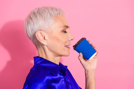 Stylish businesswoman with short white hair enjoying a coffee in vibrant attire against a pink background demonstrating relaxation.の写真素材