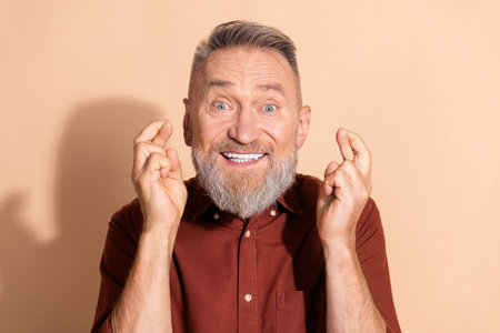 Smiling elderly man with crossed fingers, expressing hope and excitement against a beige background