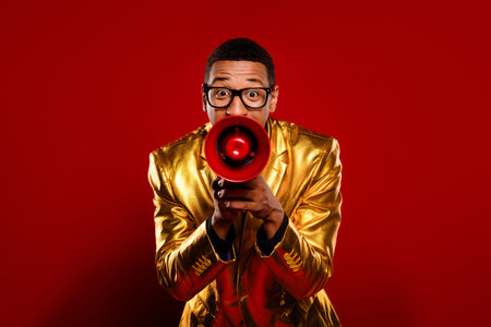 Energetic young man in a shiny gold jacket holding a red megaphone against a vibrant red background, exuding confidence and styleの写真素材